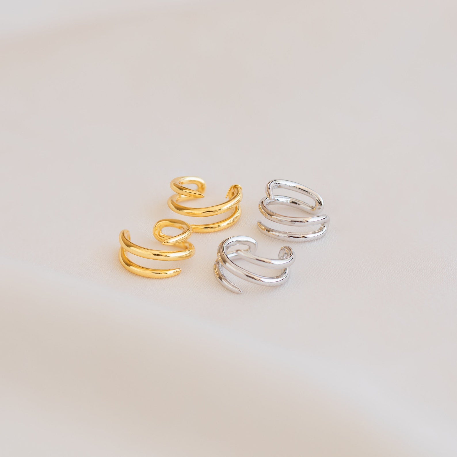 Two pairs of Claw Ear Cuffs, featuring minimalist designs in gold and silver, are displayed on a soft beige surface.