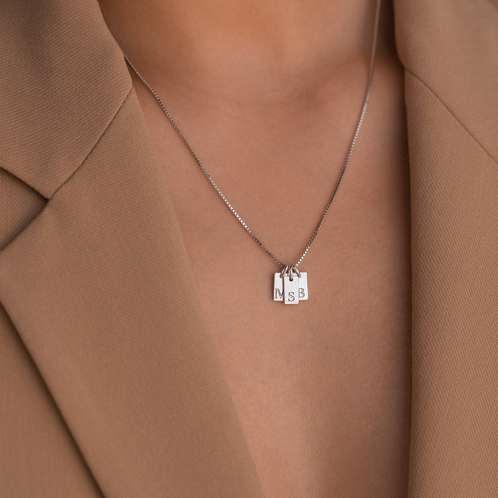 A close-up of the Mini Initial Tag Necklace with N, S, and B letter pendants, paired with a tan blazer—an elegant personalized accessory and an ideal Christmas gift.