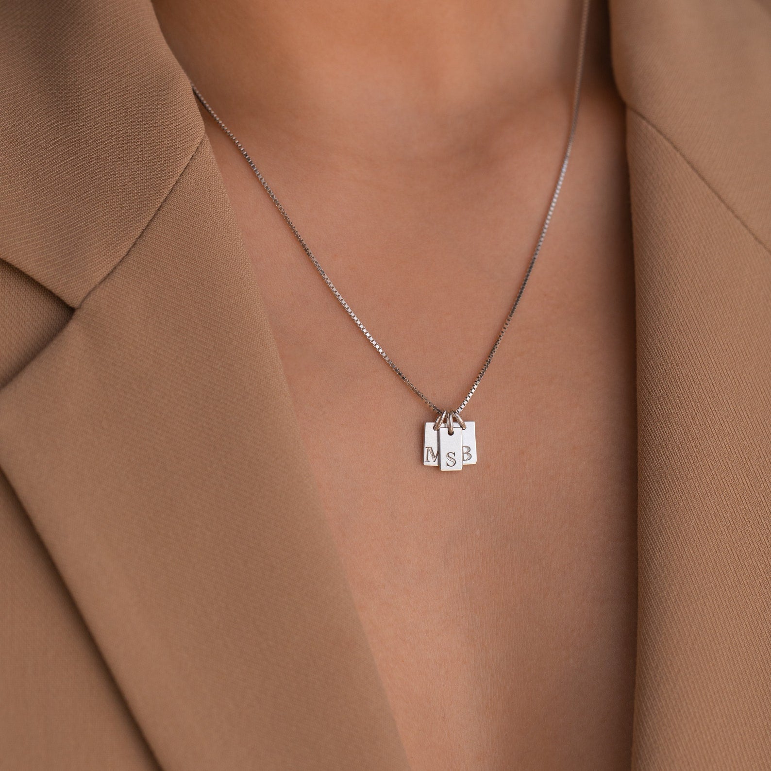 A close-up of the Mini Initial Tag Necklace with N, S, and B letter pendants, paired with a tan blazer—an elegant personalized accessory and an ideal Christmas gift.