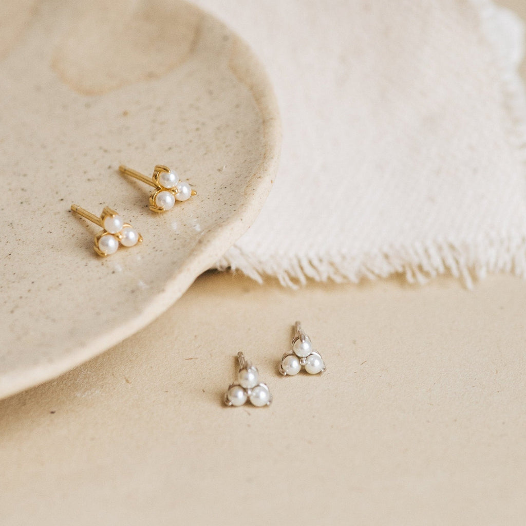 The Maud Pearl Studs, available in gold and silver, are displayed on a beige ceramic plate and fabric—luxury earrings that make a timeless gift.