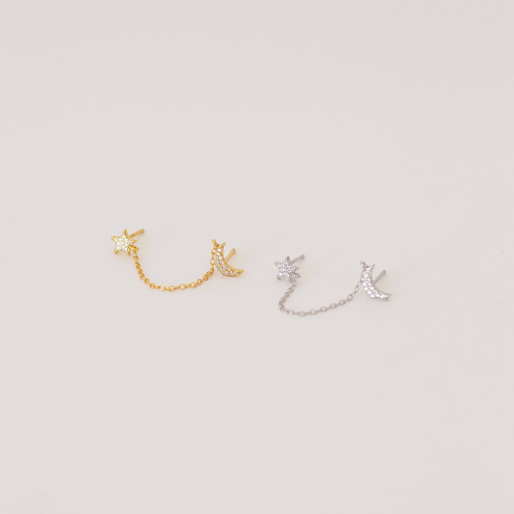The Luna Moon & Star Chain Earrings feature two pairs—one gold, one silver—linked by delicate chains, ideal for adding celestial style to your cartilage piercing. Shown on a light background.