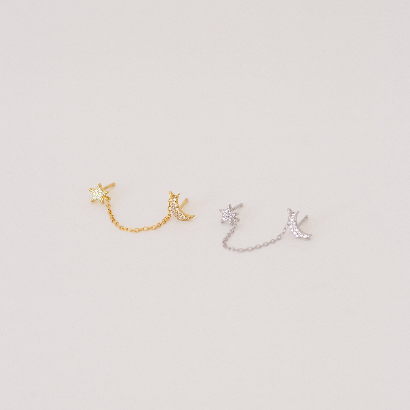 The Luna Moon & Star Chain Earrings feature two pairs—one gold, one silver—linked by delicate chains, ideal for adding celestial style to your cartilage piercing. Shown on a light background.