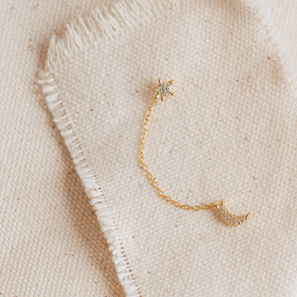 Luna Moon & Star Chain Earrings feature a gold chain with a star on one end and a crescent moon on the other, perfect for cartilage piercings and displayed on beige fabric.