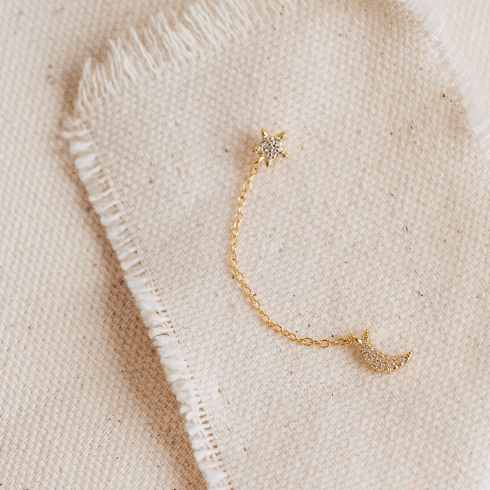 Luna Moon & Star Chain Earrings feature a gold chain with a star on one end and a crescent moon on the other, perfect for cartilage piercings and displayed on beige fabric.