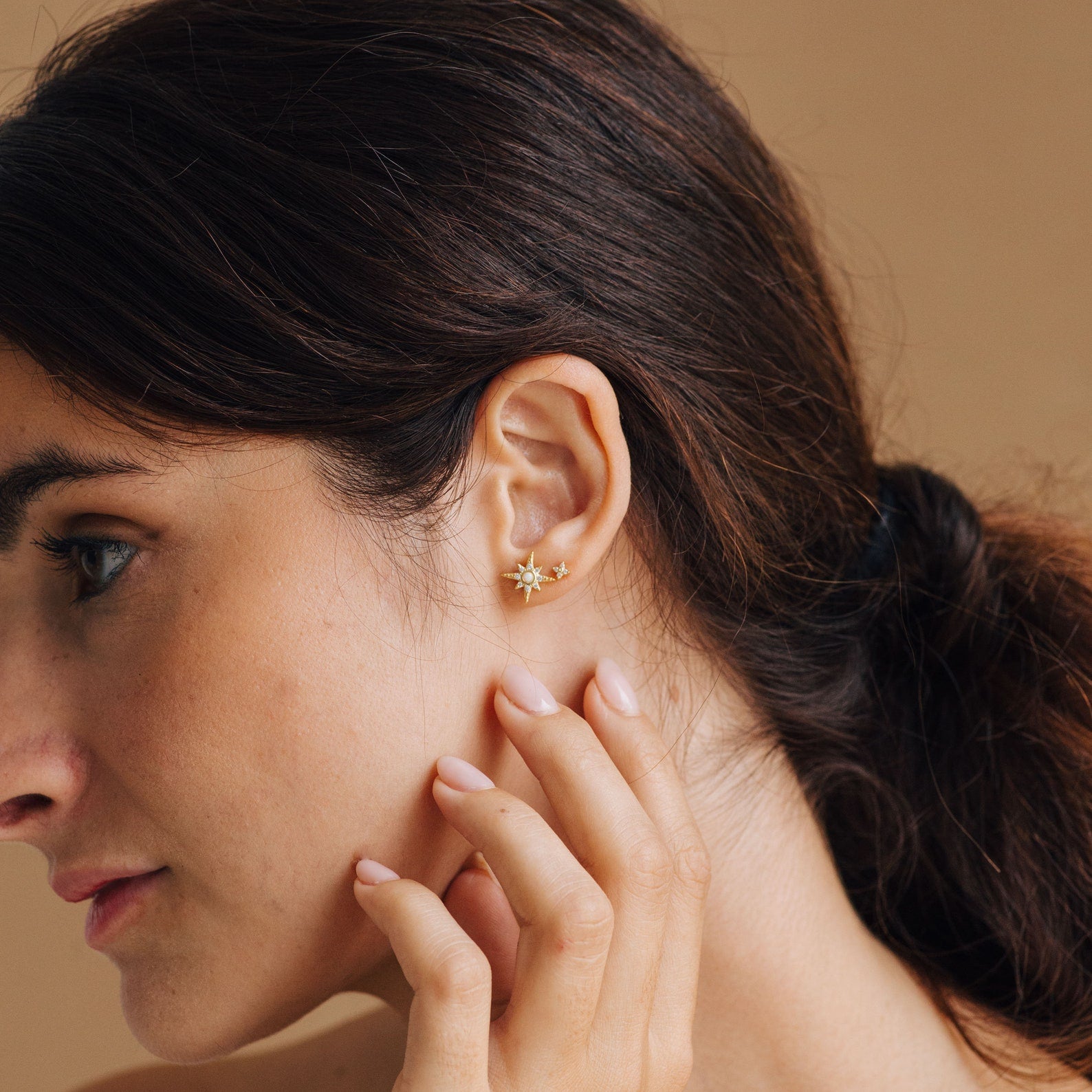 A woman with her hair tied back touches her ear, showcasing the gold Starburst Studs against a neutral background.