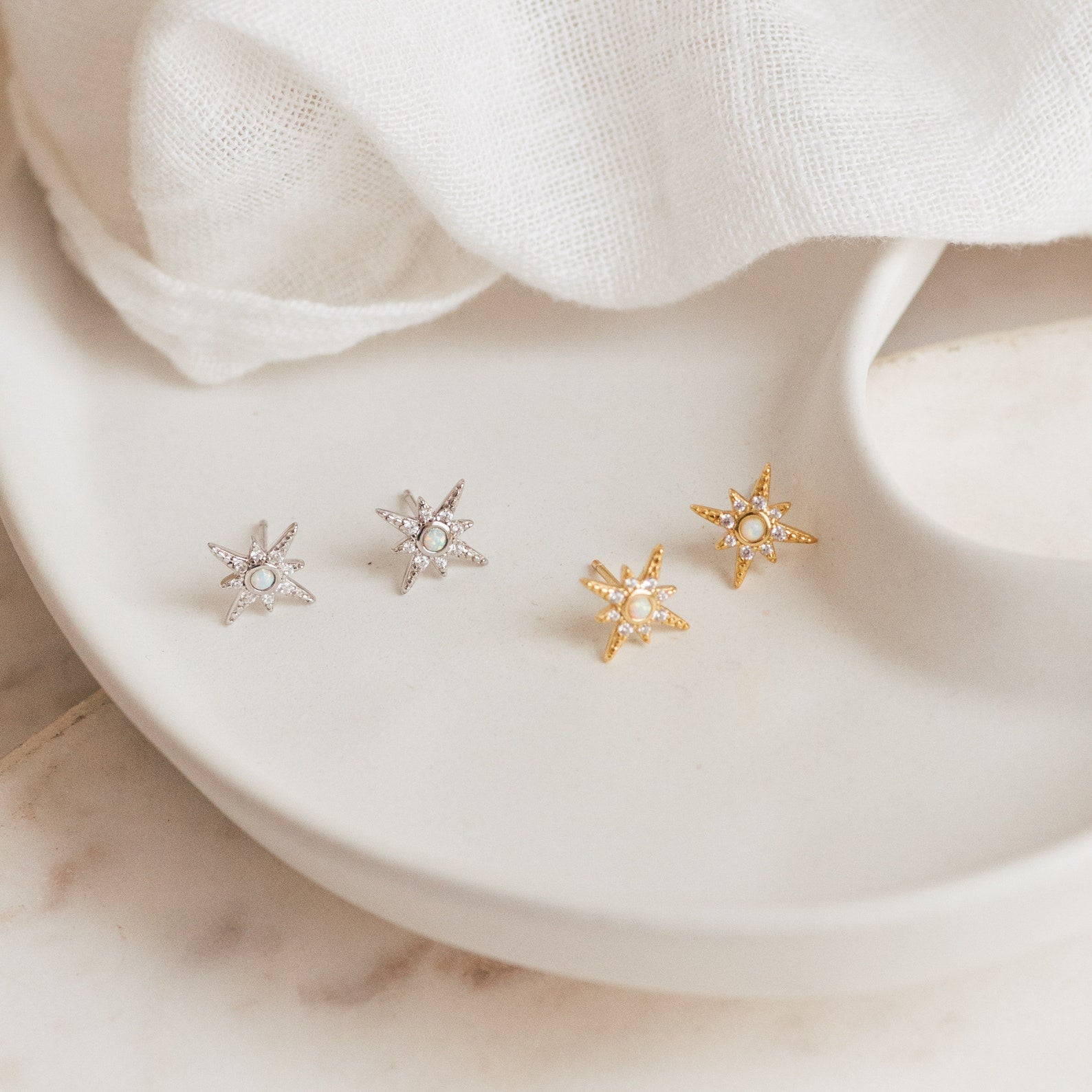 Two pairs of Starburst Studs, in silver and gold, are displayed on a white ceramic dish—perfect for adding celestial sparkle to your earring collection.