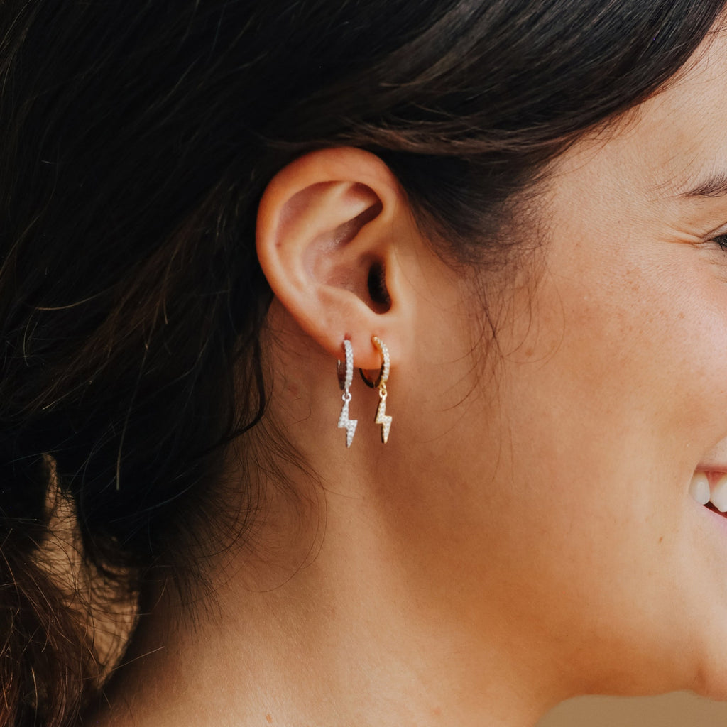 A woman wears two Striker Pave Huggies—small, trendy earrings with lightning bolt charms—in her ear, one gold and one silver.