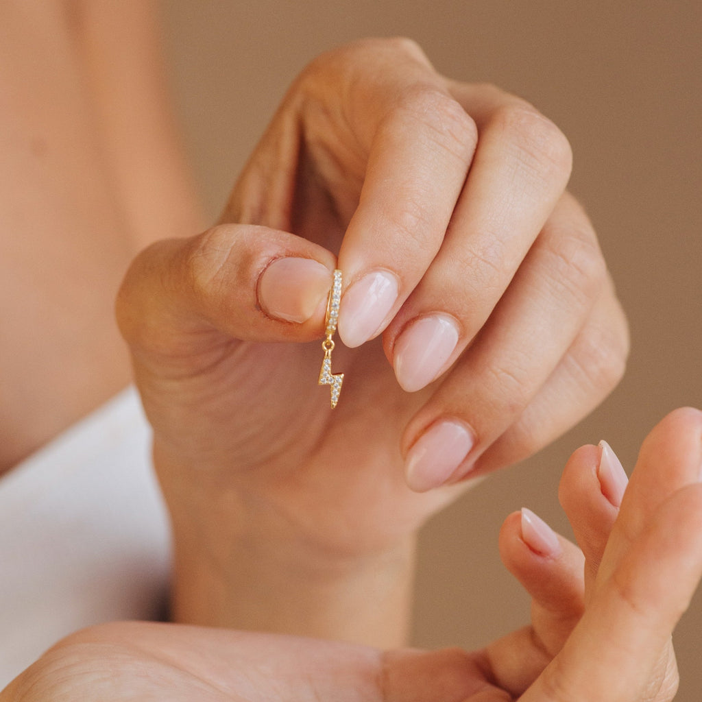A hand with manicured nails holds a gold Striker Pave Huggie earring in soft lighting, showcasing these stylish, trendy earrings.