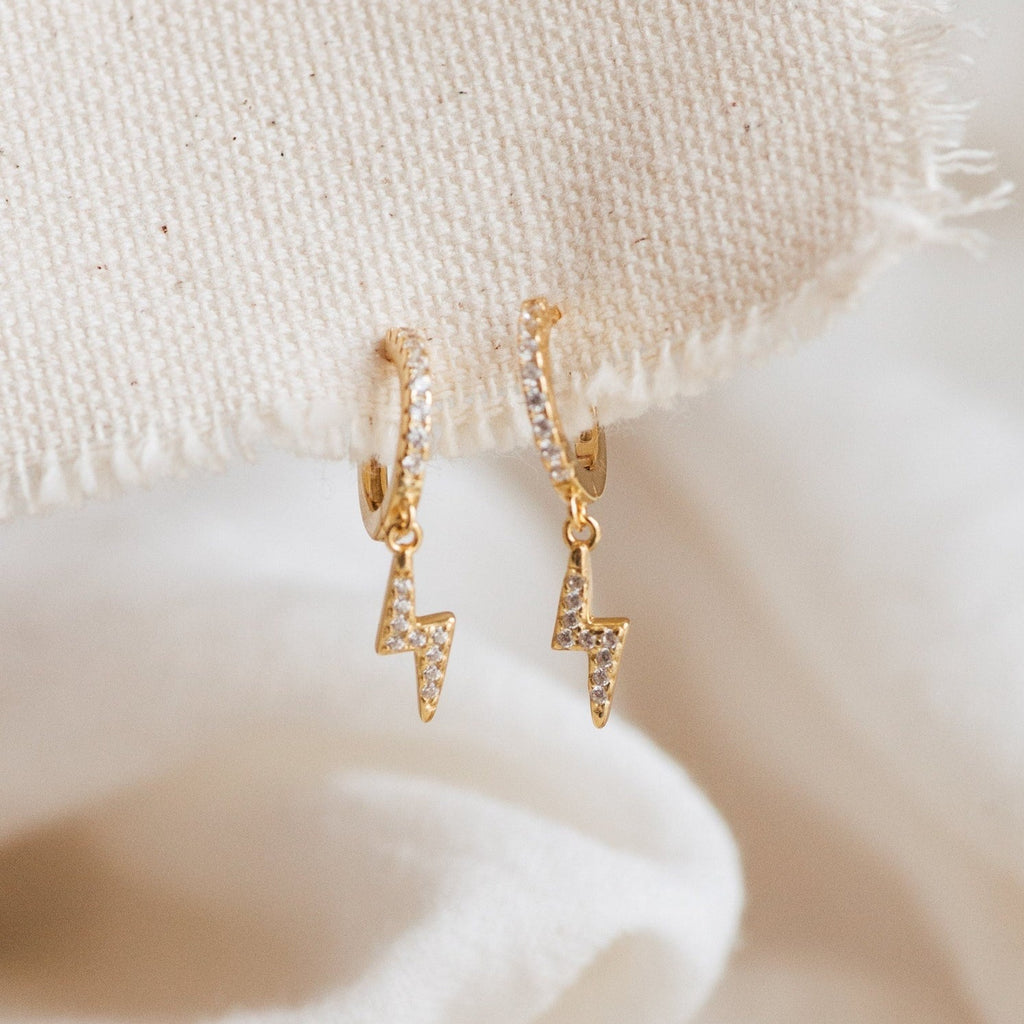 The Striker Pave Huggies feature gold hoops with lightning bolt charms and clear stone accents. These trendy earrings bring sparkle and edge to any outfit, hanging elegantly for a stylish statement.