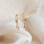 The Striker Pave Huggies feature gold hoops with lightning bolt charms and clear stone accents. These trendy earrings bring sparkle and edge to any outfit, hanging elegantly for a stylish statement.