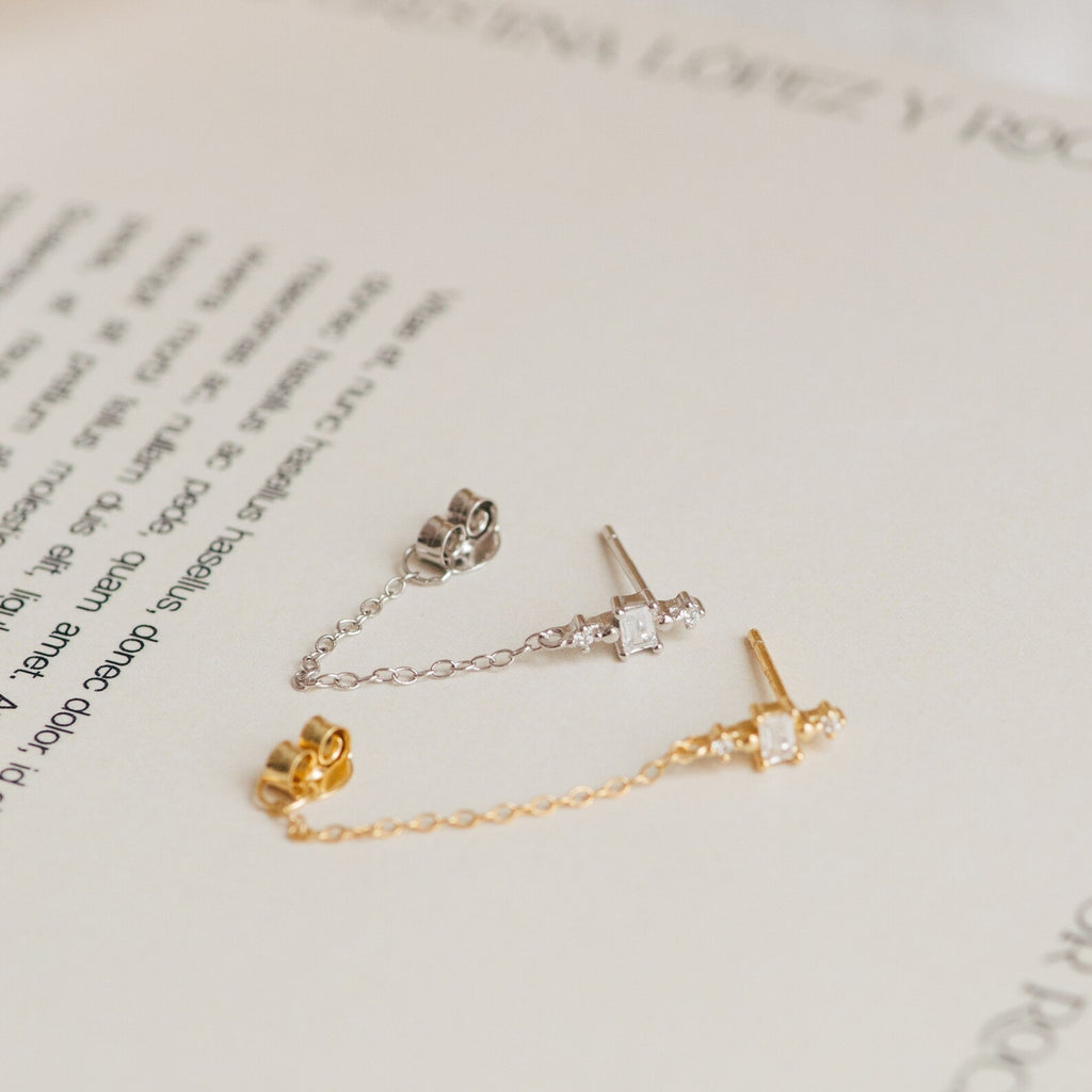 The Florence Diamond Chain Earrings feature two delicate studs—one silver and one gold—with crystal accents and fine chains, beautifully displayed on an open book.