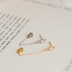 The Florence Diamond Chain Earrings feature two delicate studs—one silver and one gold—with crystal accents and fine chains, beautifully displayed on an open book.