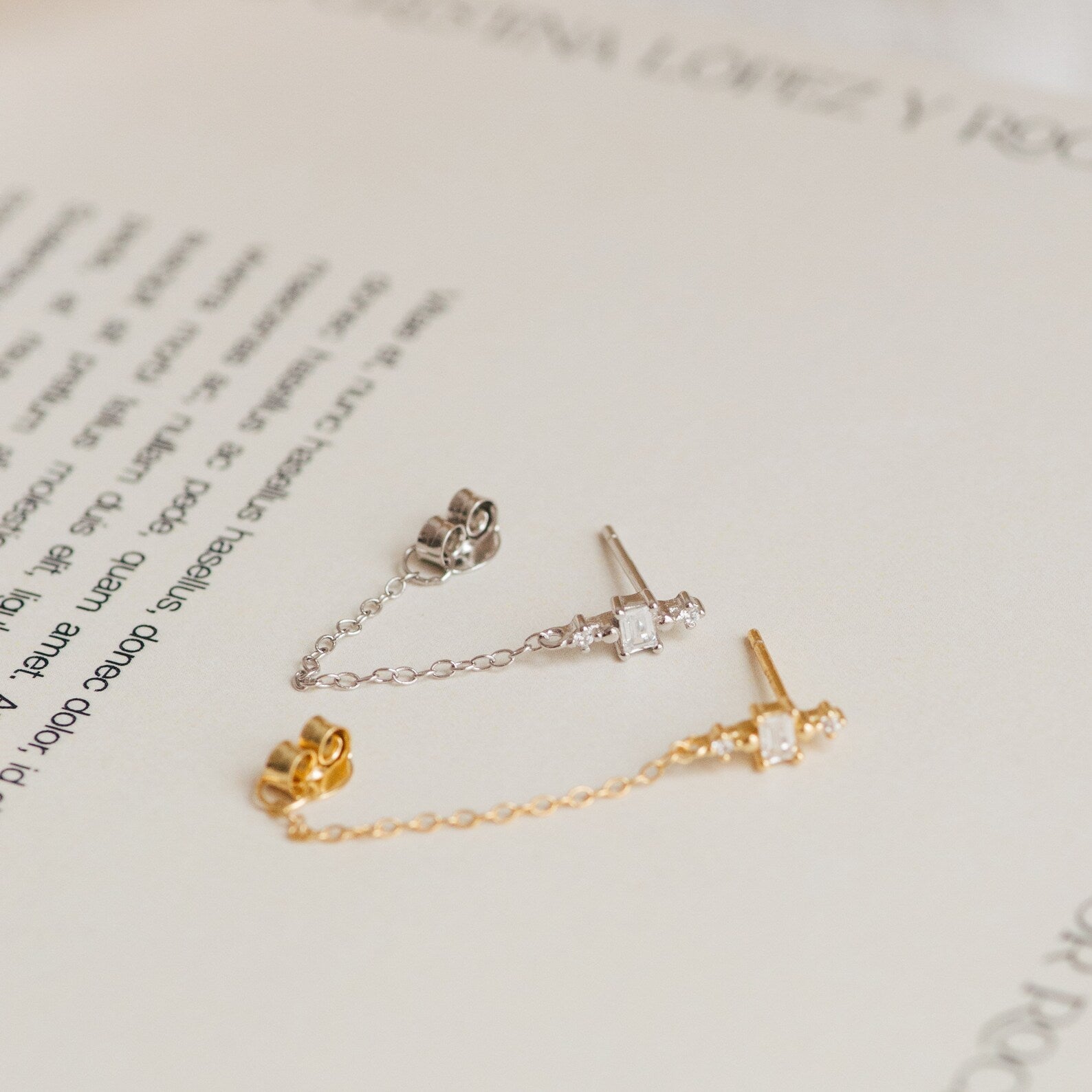 The Florence Diamond Chain Earrings feature two delicate studs—one silver and one gold—with crystal accents and fine chains, beautifully displayed on an open book.