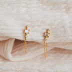 The Florence Diamond Chain Earrings, featuring delicate hanging chains and sparkling diamond accents, are displayed on a soft, cream-colored fabric.