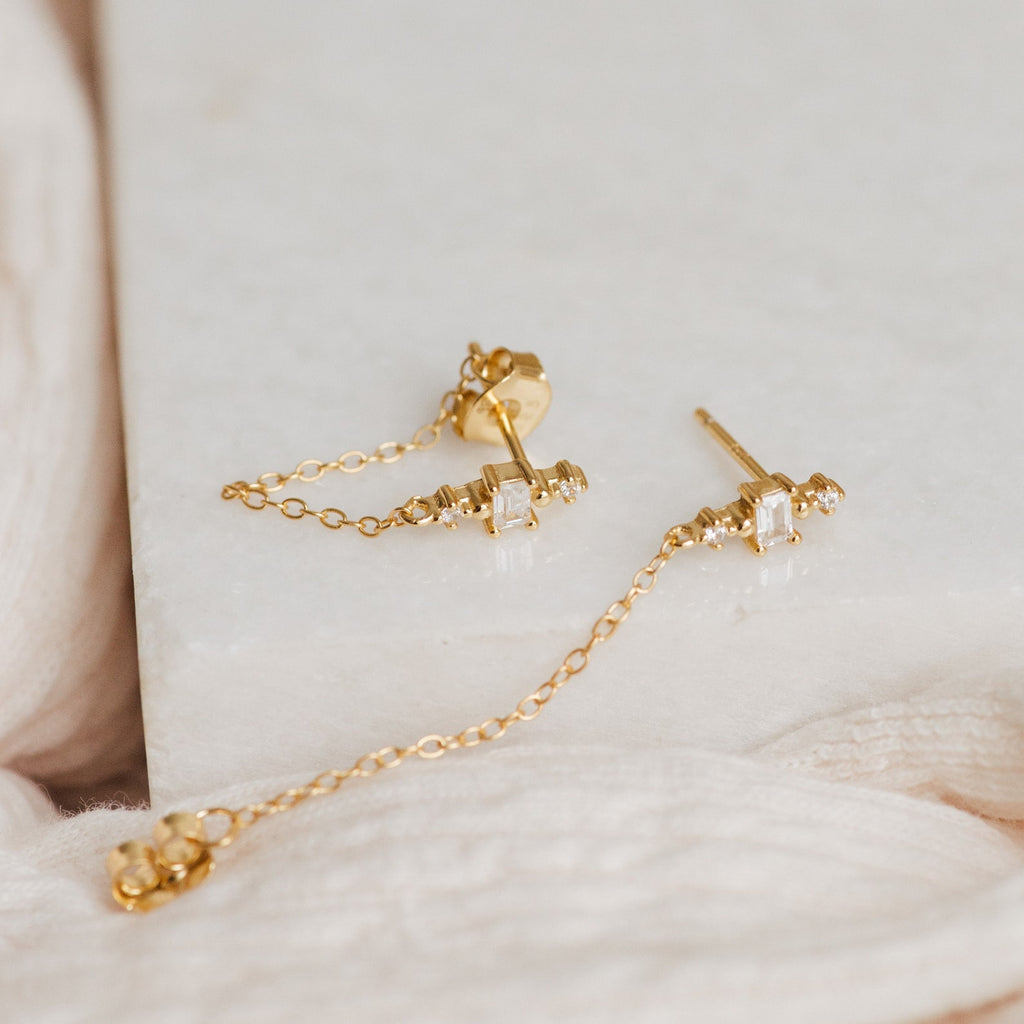 Florence Diamond Chain Earrings featuring gold studs, dangling chains, and crystal accents, displayed on marble with soft fabric.
