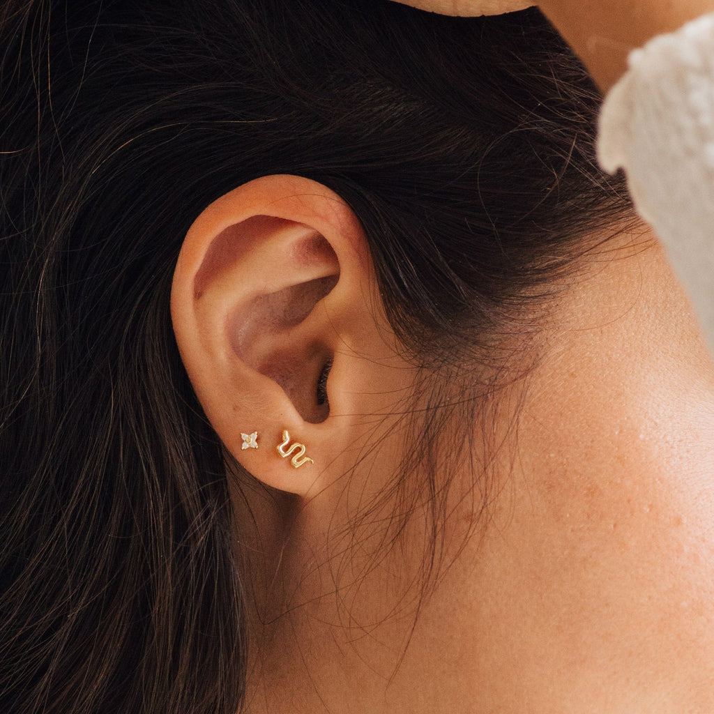 Close-up of a woman's ear adorned with dainty jewelry: a small diamond stud and gold Tiny Python Studs, perfect for those who love boho-luxe earrings.