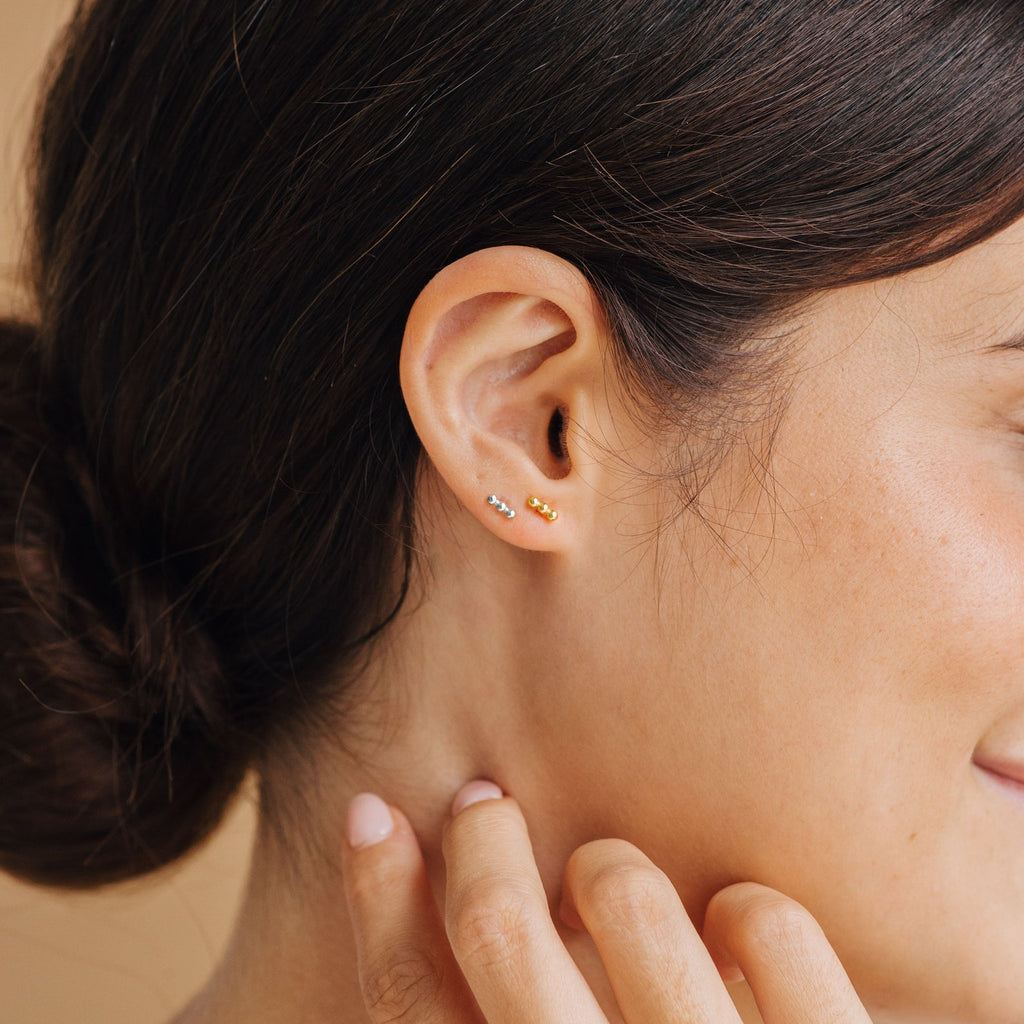 A woman with dark hair in a bun, wearing Trio Beaded Bar Studs and touching her neck, showcases a look perfect for everyday wear.