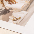 Tiny Pave Safety Pin Studs with sparkling CZ diamond accents rest on a soft beige fabric, complemented by delicate dried flowers in the corner.