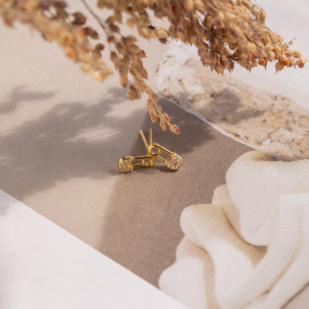 Tiny Pave Safety Pin Studs featuring small CZ crystals are displayed on a neutral background with dried flowers above.