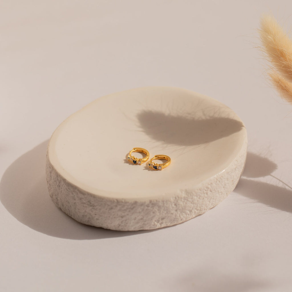 The Angelica Sapphire Huggies, gold rings set with blue gemstones, are displayed on a round, textured ceramic dish in soft natural light and paired elegantly with minimal earrings.
