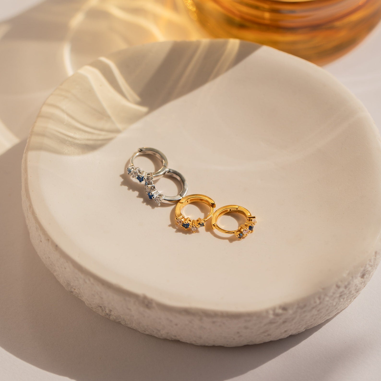 A pair of Angelica Sapphire Huggies in gold and silver are displayed on a round, off-white ceramic dish.