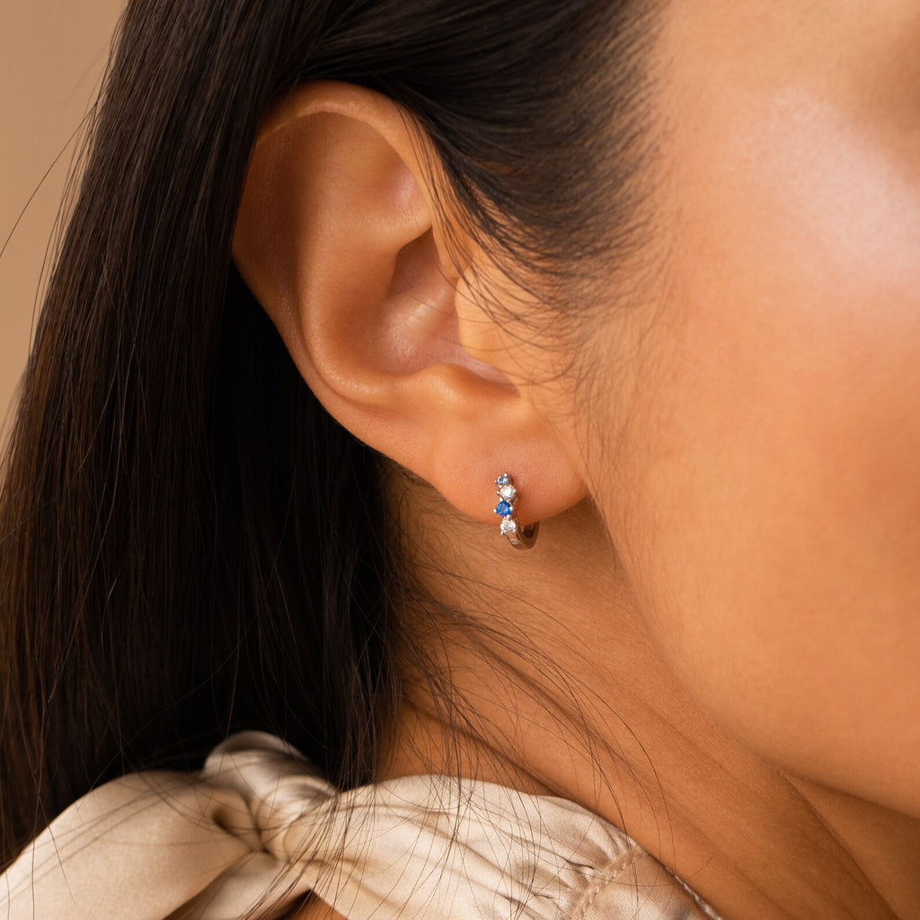 Close-up of a woman's ear wearing the Angelica Sapphire Huggies, stylish minimal hoops adorned with blue and white gemstones.