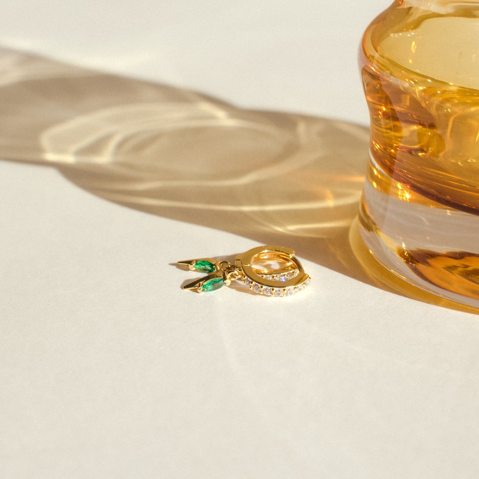 The Raine Emerald Dagger Huggies—gold hoop earrings with green and clear stones—rest beside a glass casting light on a white surface, making an elegant gift for her.