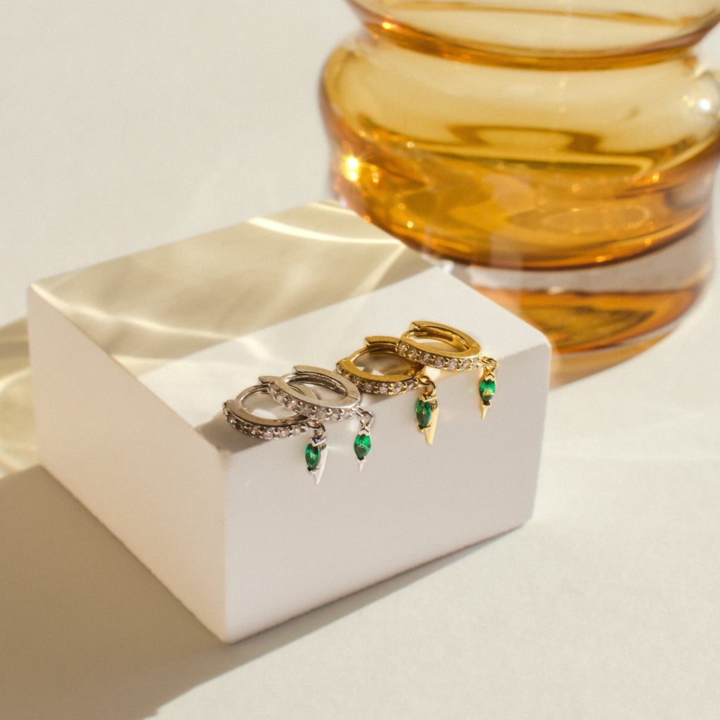 Two pairs of hoop earrings—silver and gold with green stones—are displayed on a white block near amber glass. The Raine Emerald Dagger Huggies make a perfect gift for her. Shop elegant designs, including diamond and emerald huggie earrings.