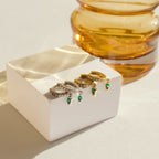 Two pairs of hoop earrings—silver and gold with green stones—are displayed on a white block near amber glass. The Raine Emerald Dagger Huggies make a perfect gift for her. Shop elegant designs, including diamond and emerald huggie earrings.