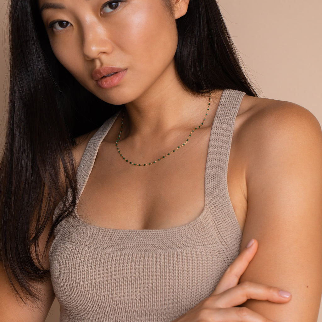 A woman in a beige knit tank top poses against a beige background, accessorized with the Emerald Station Necklace featuring delicate emerald green beads for a boho touch.