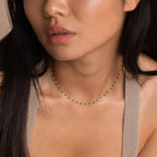 A woman wearing the Emerald Station Necklace, featuring delicate gold charms and emerald green beads, paired with a beige knit top.