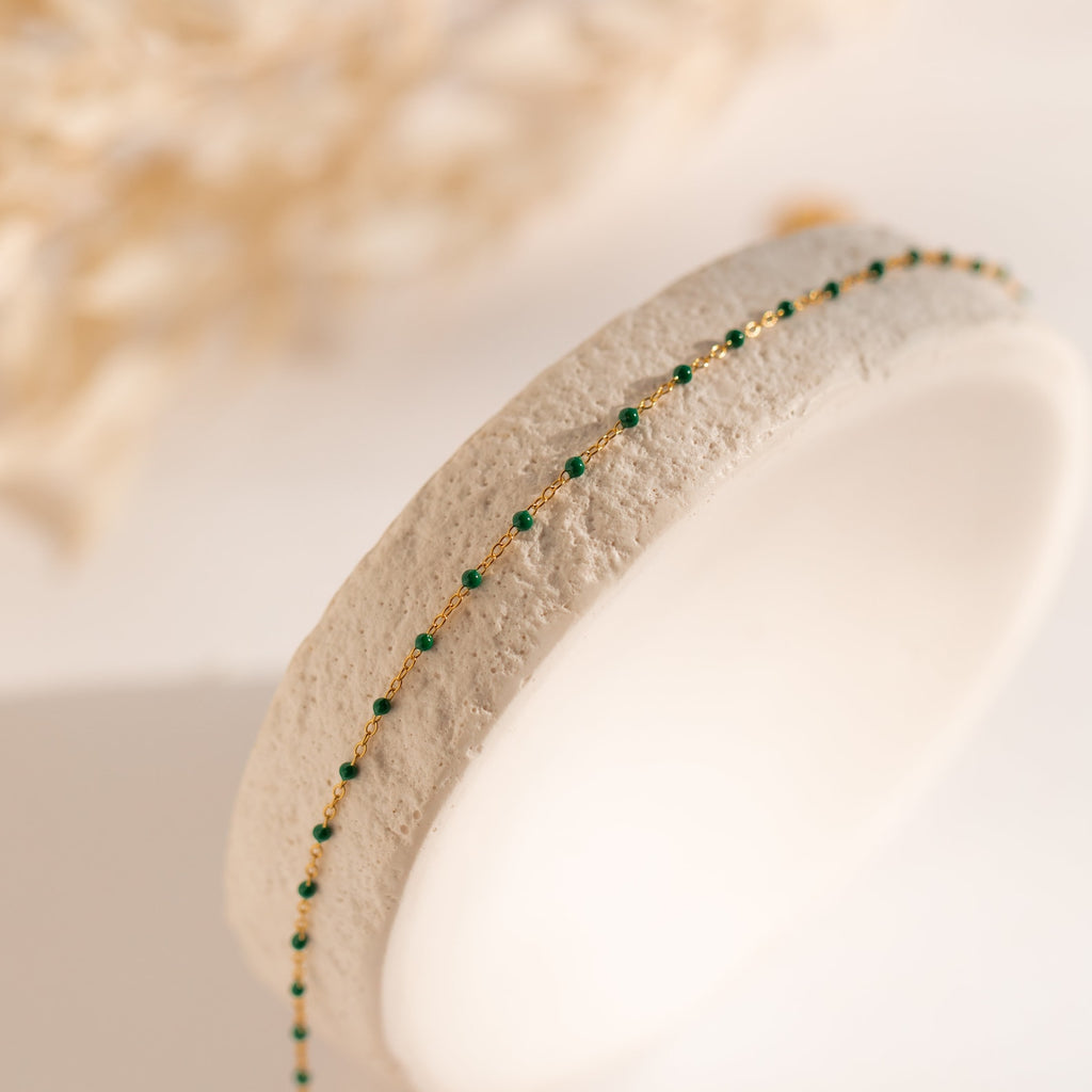 The Emerald Station Necklace features a delicate gold chain adorned with small emerald green beads, presented on a textured white stand.