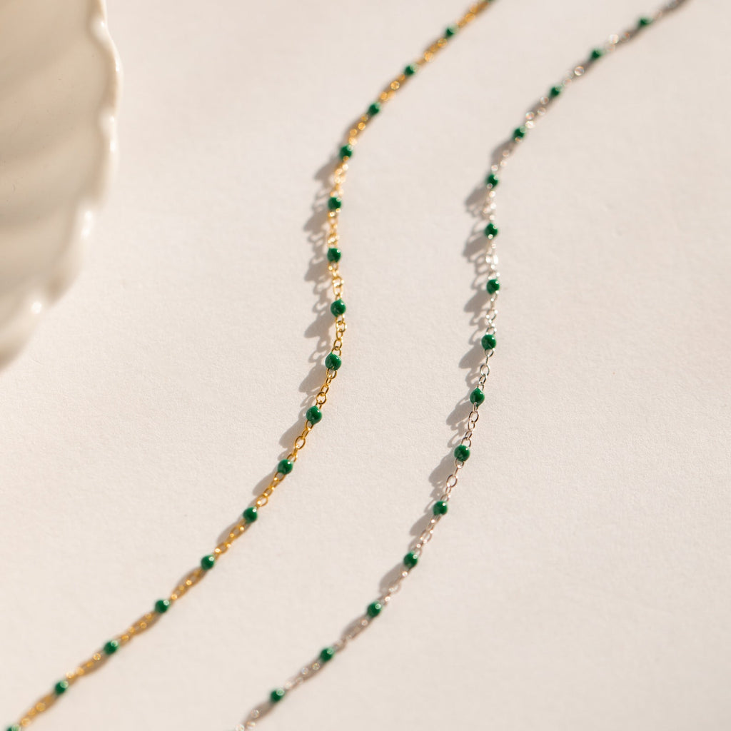 The Emerald Station Necklace, featuring delicate chains with emerald green beads in both gold and silver, adds beaded boho charm—beautifully displayed on a white surface.