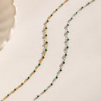 The Emerald Station Necklace, featuring delicate chains with emerald green beads in both gold and silver, adds beaded boho charm—beautifully displayed on a white surface.