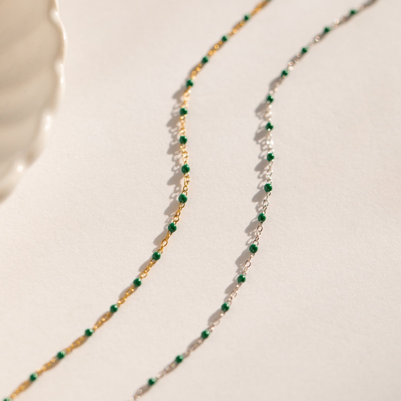 The Emerald Station Necklace, featuring delicate chains with emerald green beads in both gold and silver, adds beaded boho charm—beautifully displayed on a white surface.