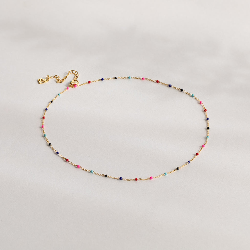 The Rainbow Station Necklace features a delicate gold chain with small, colorful beads evenly spaced along its length, shown on a light background.