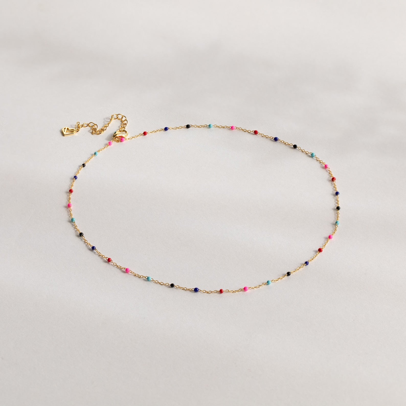 The Rainbow Station Necklace features a delicate gold chain with small, colorful beads evenly spaced along its length, shown on a light background.