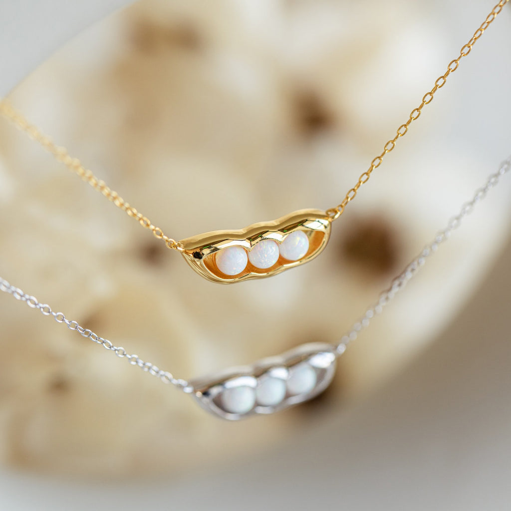 The Peas in a Pod Necklace features three white pearls nestled in a gold and silver pod, resting on a soft beige background—an elegant piece symbolizing special bonds.
