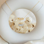 The Peas in a Pod Necklace, available in gold and silver, is shown on a white circular stand—meaningful jewelry that celebrates the connection between loved ones.