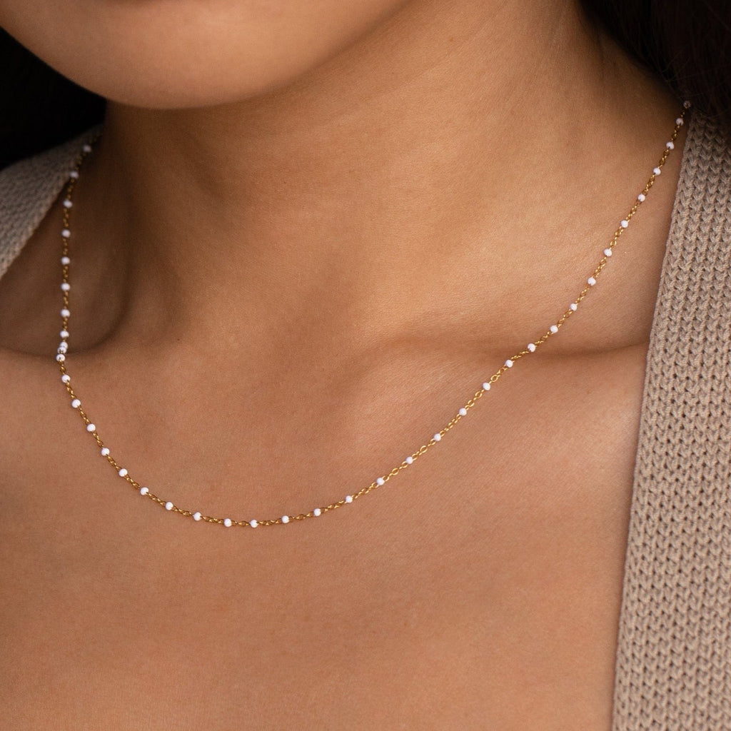 Close-up of a woman wearing the White Station Necklace, a delicate gold chain with dainty white beads, paired with a beige knit sweater.