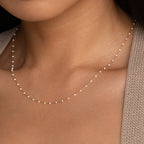 Close-up of a woman wearing the White Station Necklace, a delicate gold chain with dainty white beads, paired with a beige knit sweater.