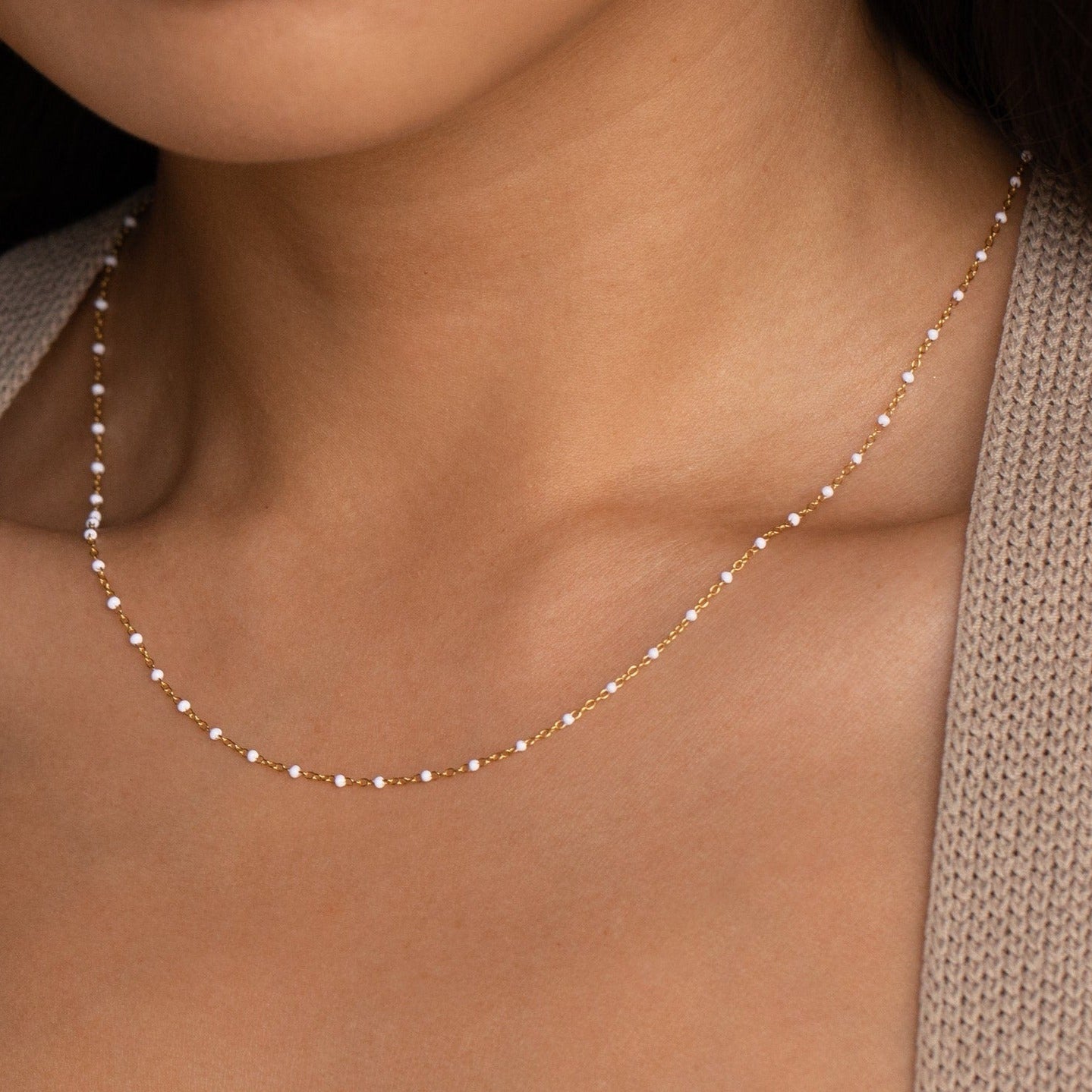 Close-up of a woman wearing the White Station Necklace, a delicate gold chain with dainty white beads, paired with a beige knit sweater.