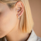 Close-up of a person with short blonde hair wearing the Astra Moon & Star Huggies: opal-studded hoops featuring delicate dangling star and moon charms.