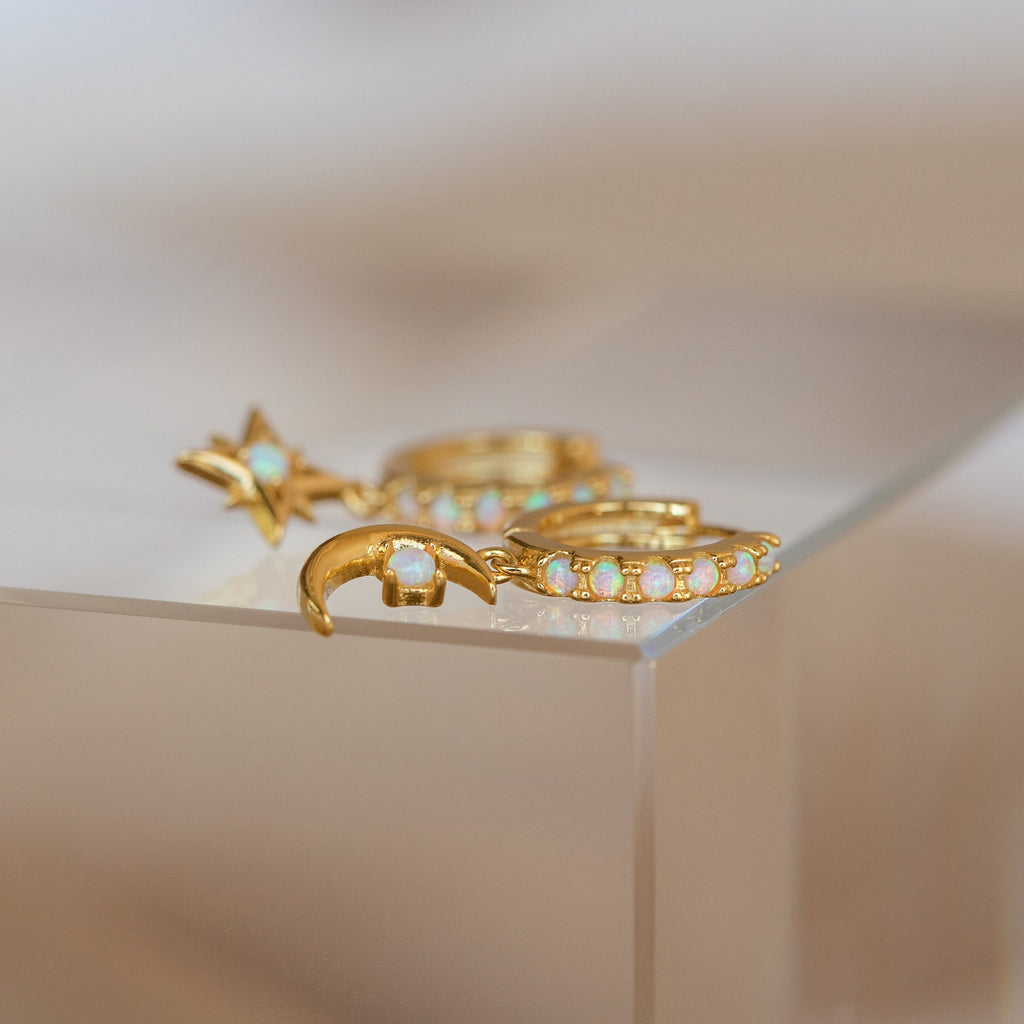 Astra Moon & Star Huggies: Gold earrings with opal accents in star, crescent moon, and opal-studded huggie hoop designs rest elegantly on a glass surface.
