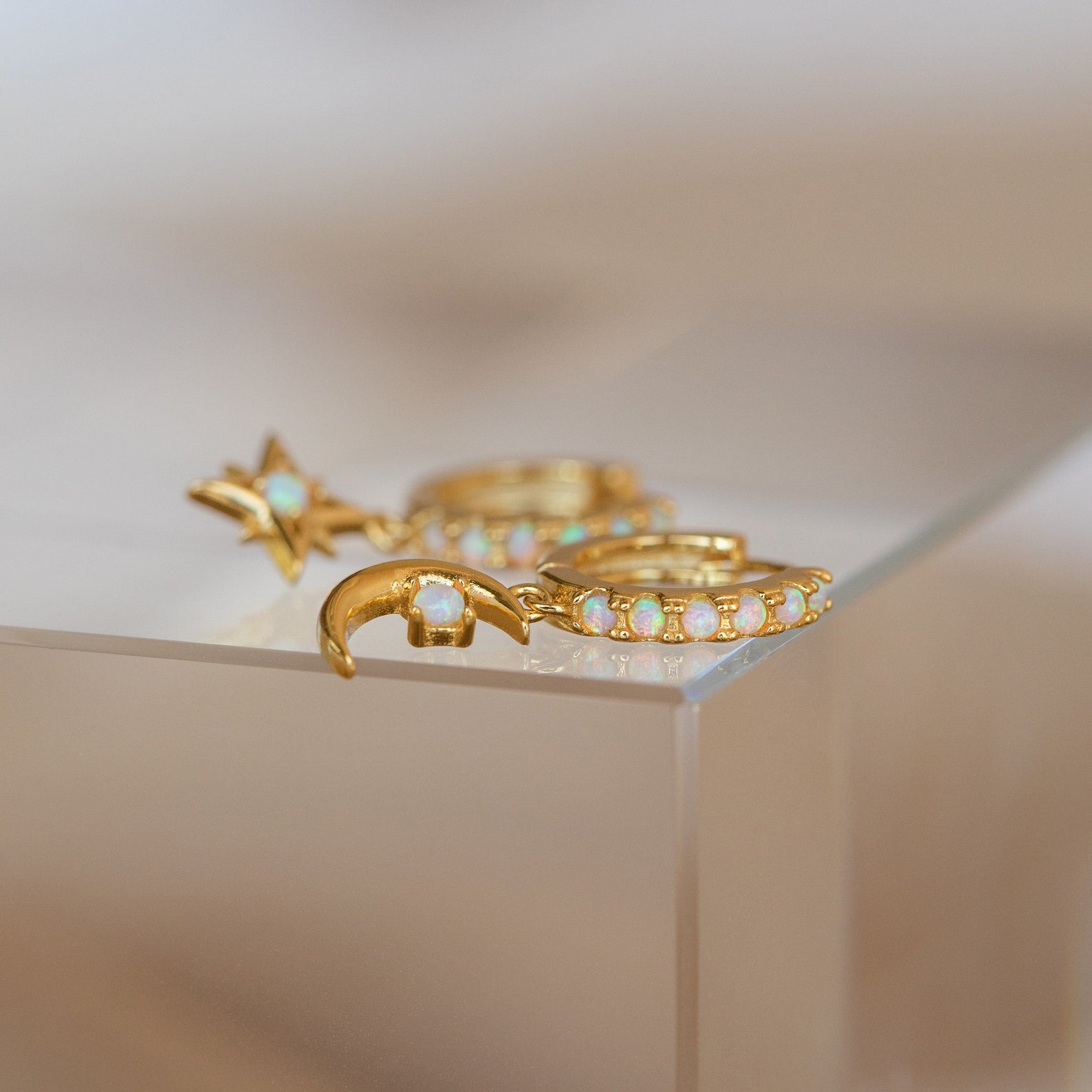 Astra Moon & Star Huggies: Gold earrings with opal accents in star, crescent moon, and opal-studded huggie hoop designs rest elegantly on a glass surface.