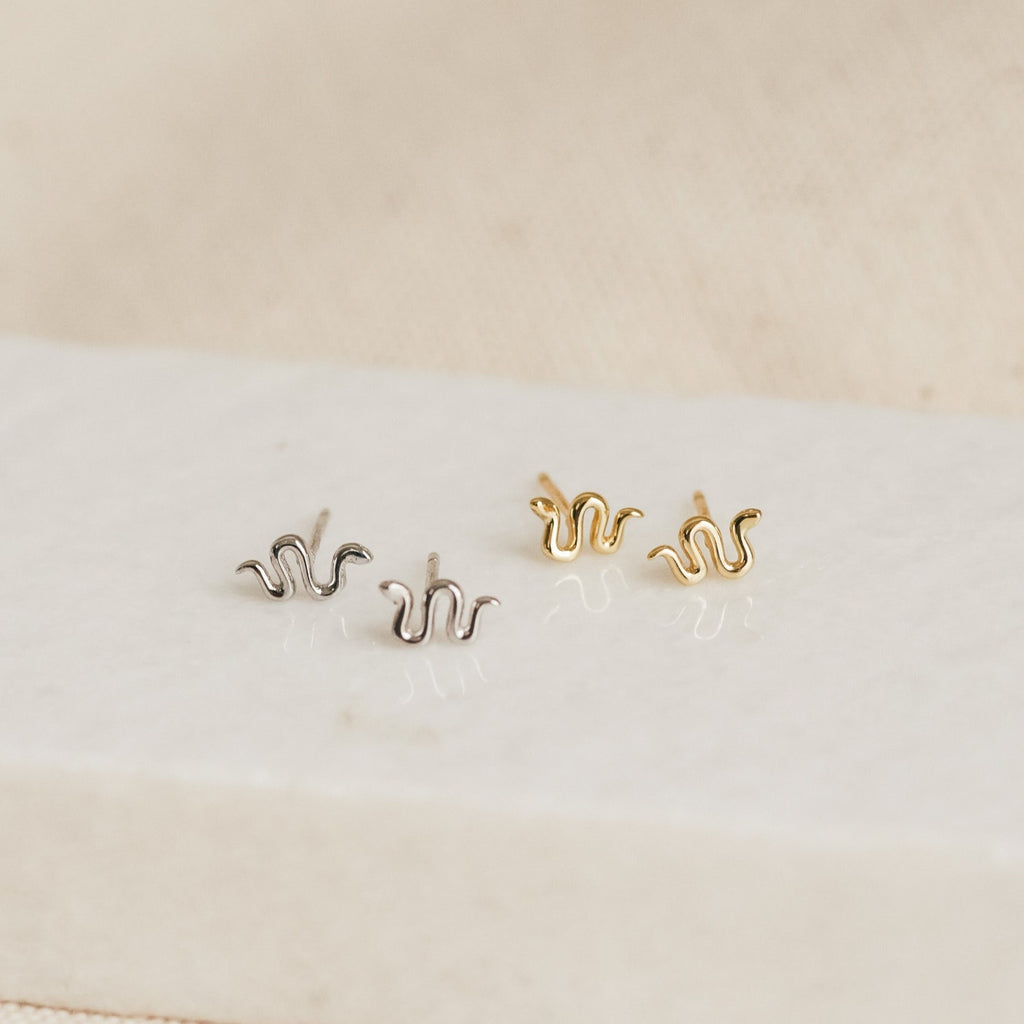The Tiny Python Studs, featuring one silver and one gold wavy pair, are displayed on a white rectangular surface—ideal for those who love dainty or boho-luxe jewelry.