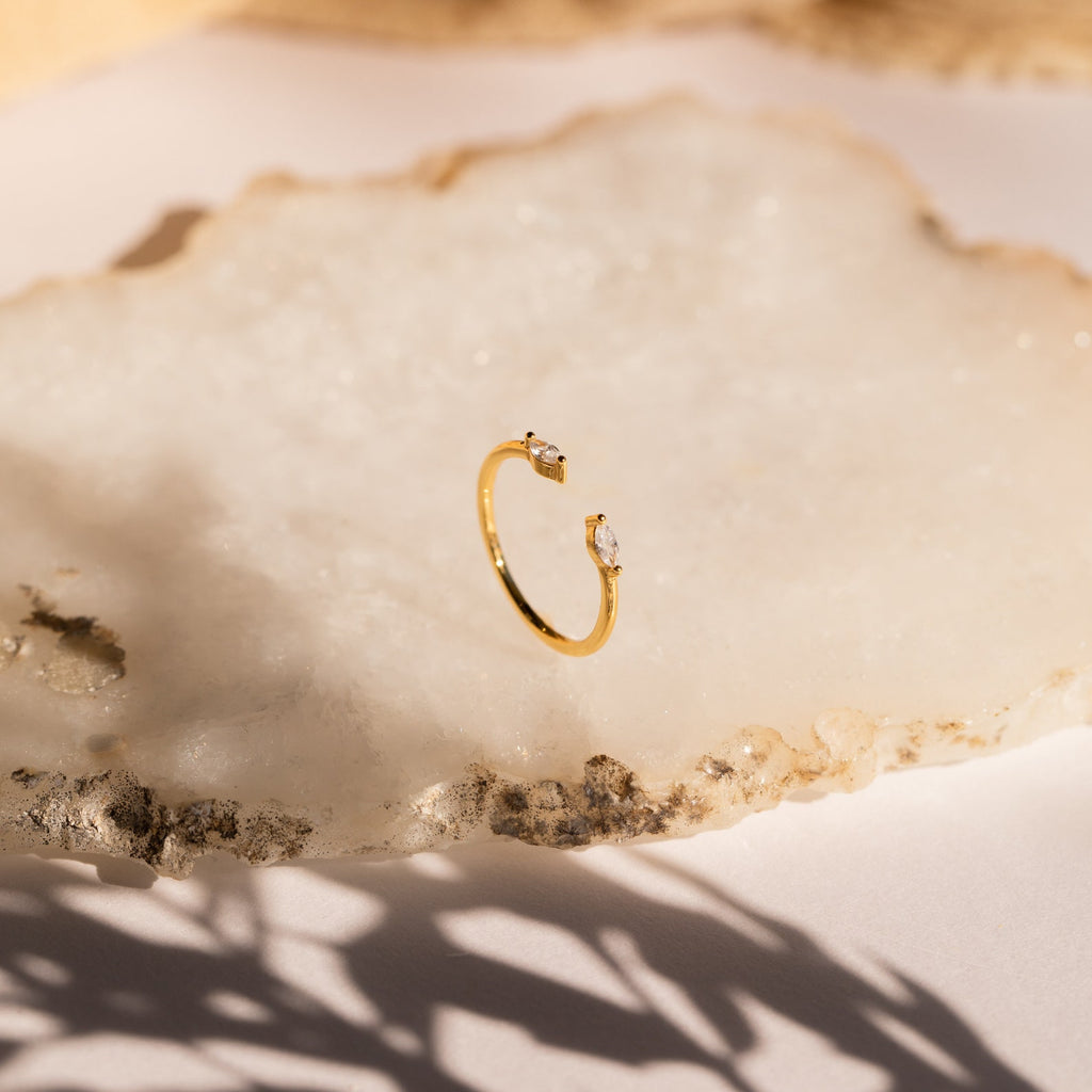The Celeste Duo Marquise Ring, a gold open ring featuring two gemstones, is displayed on a textured, light-colored stone surface.