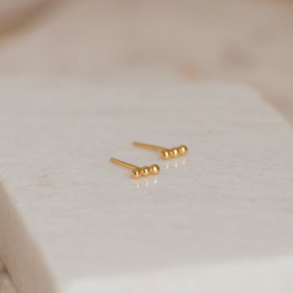 Minimalist and perfect for daily wear, the Trio Beaded Bar Studs feature three polished gold beads, shown on a textured white background.