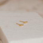Minimalist and perfect for daily wear, the Trio Beaded Bar Studs feature three polished gold beads, shown on a textured white background.