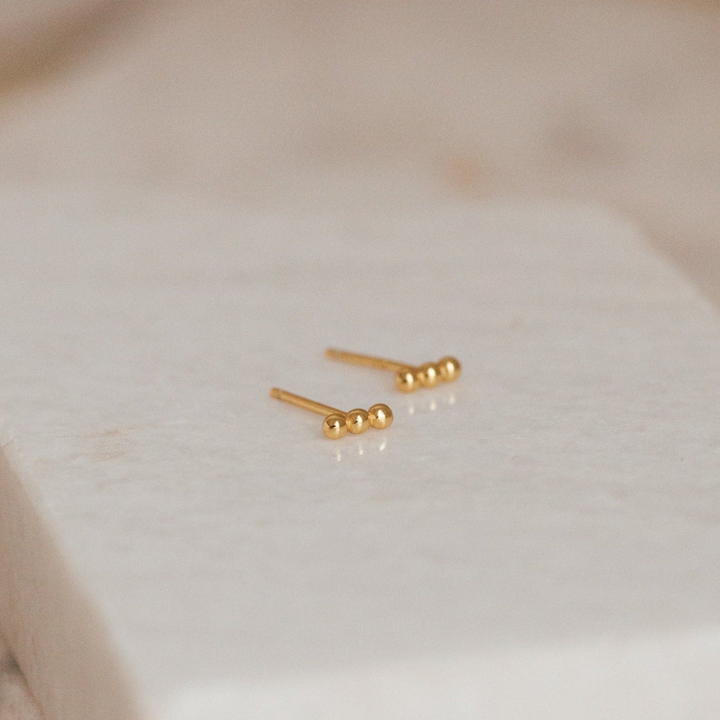 Minimalist and perfect for daily wear, the Trio Beaded Bar Studs feature three polished gold beads, shown on a textured white background.